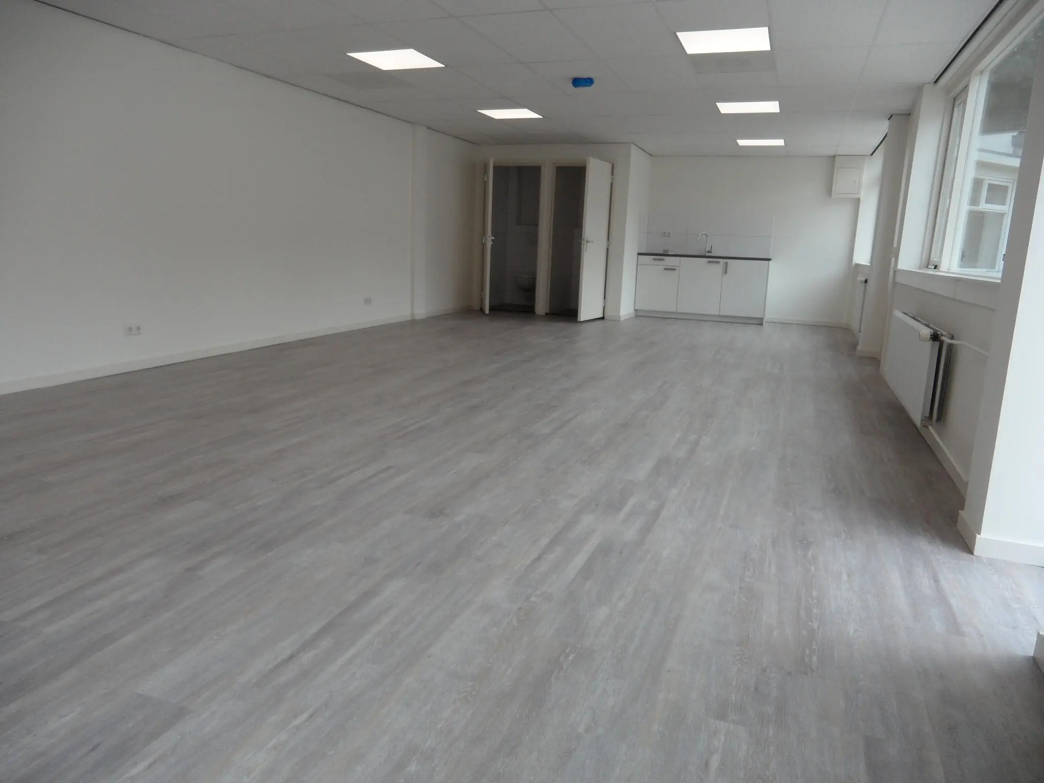 Lege, lichte ruimte met grijze laminaatvloer, systeemplafond en een kitchenette aan de achterzijde, gelegen aan de Jacques Perkstraat.